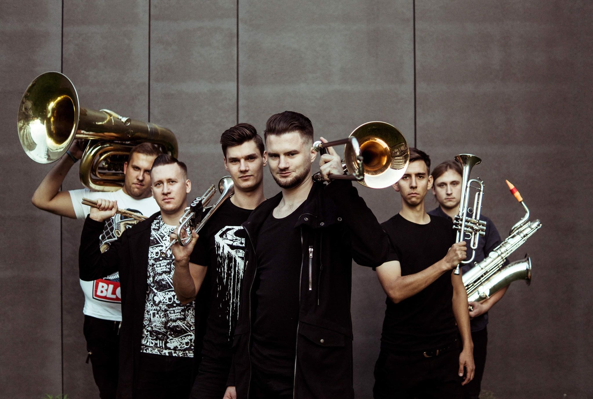 Sześcioosobowy zespół muzyczny pozuje z instrumentami dętymi na tle szarej ściany. Widoczna tuba, puzony i saksofon.