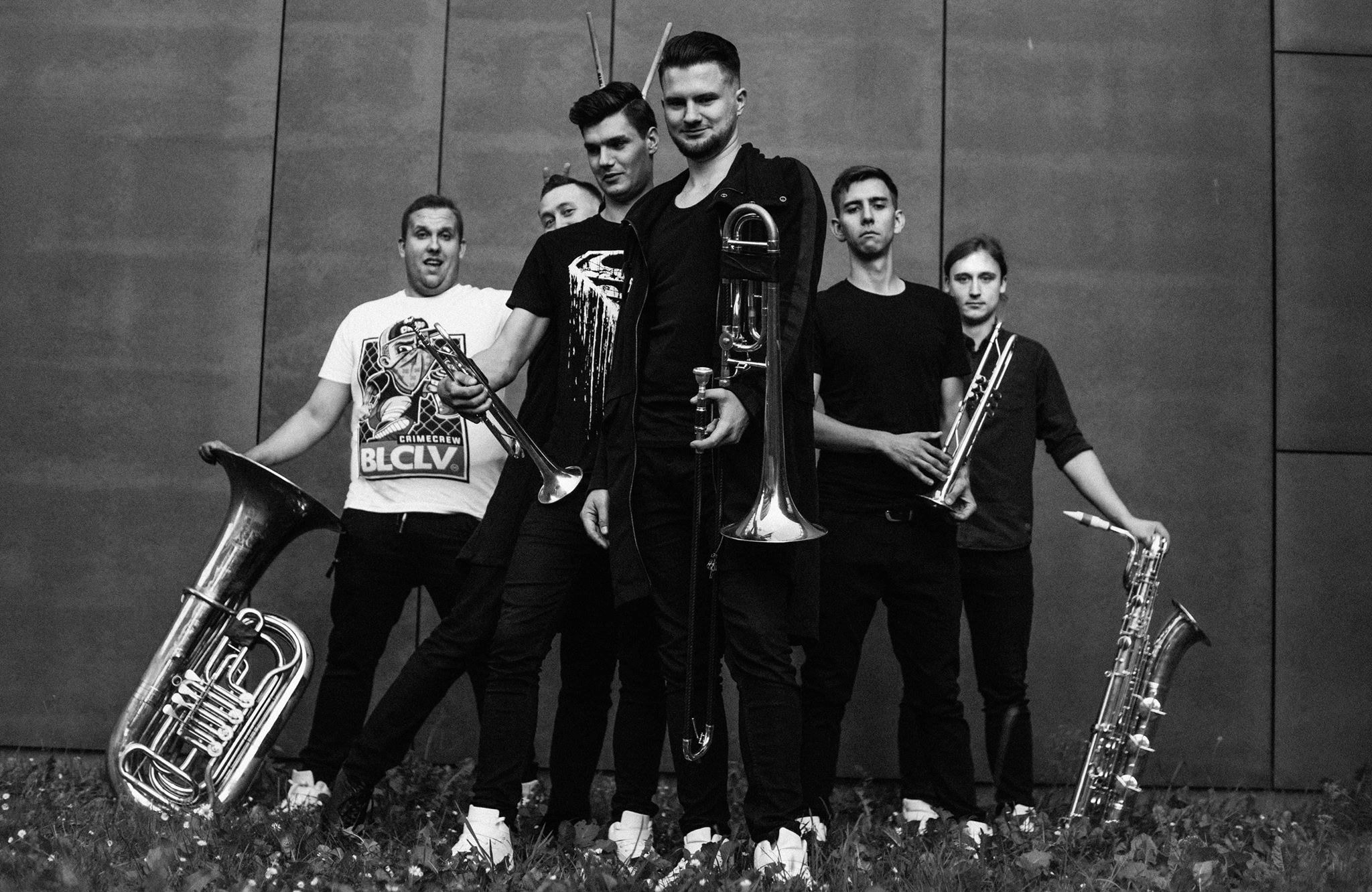 Czarno-białe zdjęcie zespołu muzycznego z Krakowa, prezentującego instrumenty dęte: tubę, trąbki i saksofon, na tle nowoczesnej ściany i trawy.