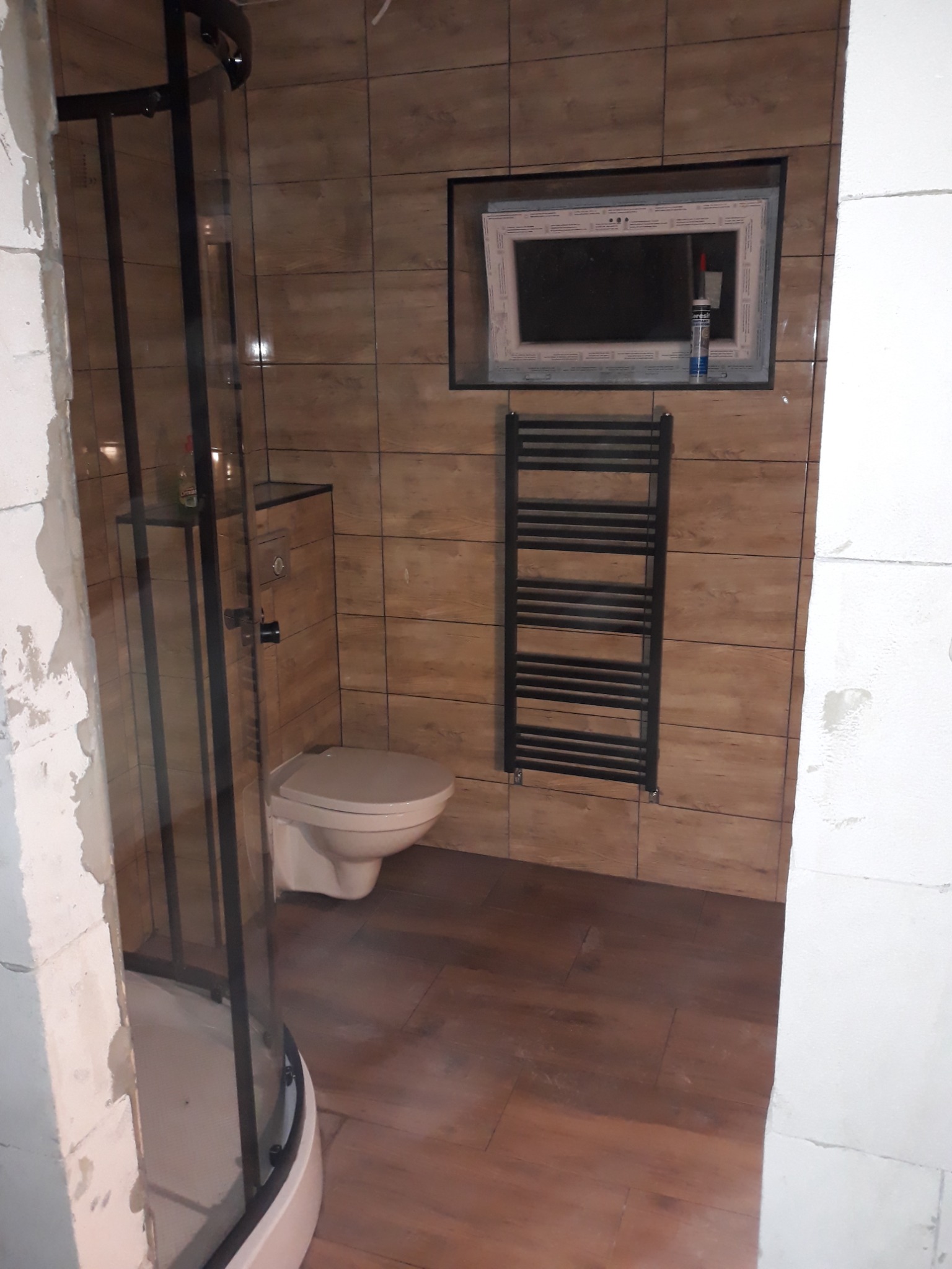 Niedokończona łazienka z brązowymi płytkami imitującymi drewno, widoczna kabina prysznicowa z czarnymi profilami, wisząca toaleta, grzejnik drabinkowy i okno z tubą silikonu na parapecie.