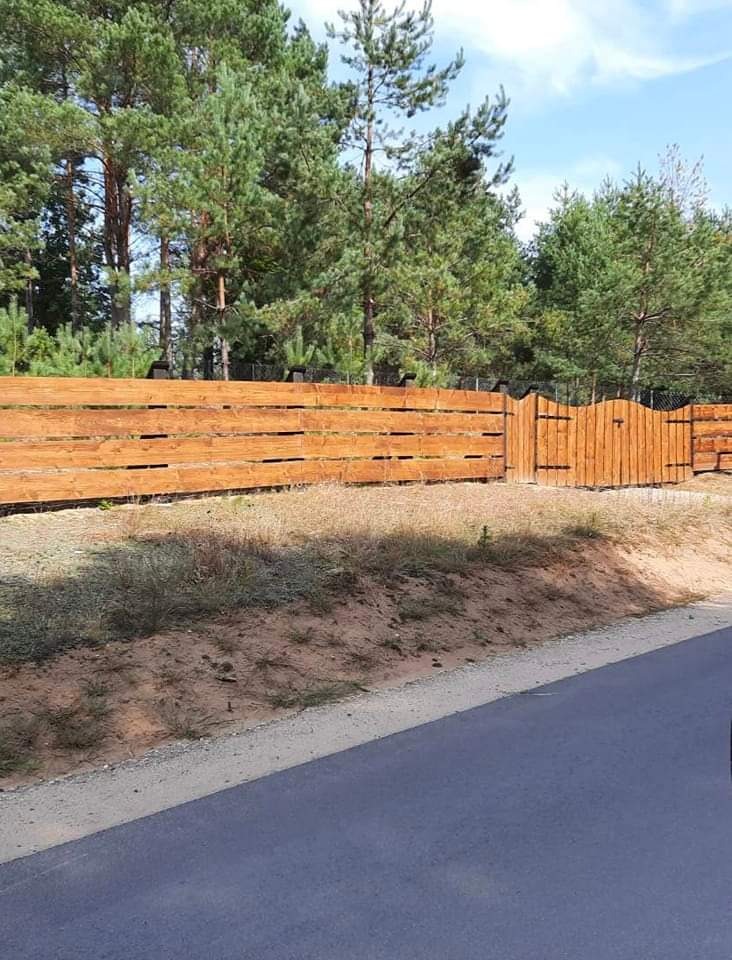 Nowo postawione, drewniane ogrodzenie panelowe z bramką, pomalowane na brązowo, z widocznymi słojami drewna, otoczone drzewami iglastymi i trawą, widziane z perspektywy ulicy.