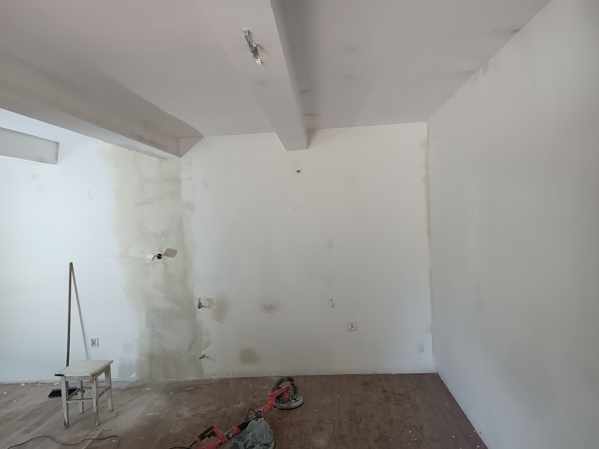 Pokój w trakcie szpachlowania ścian, widoczna szpachelka oparta o ścianę, szlifierka do gładzi leżąca na podłodze z panelami, krzesło malarskie obok ściany z widocznymi puszkami elektrycznymi.