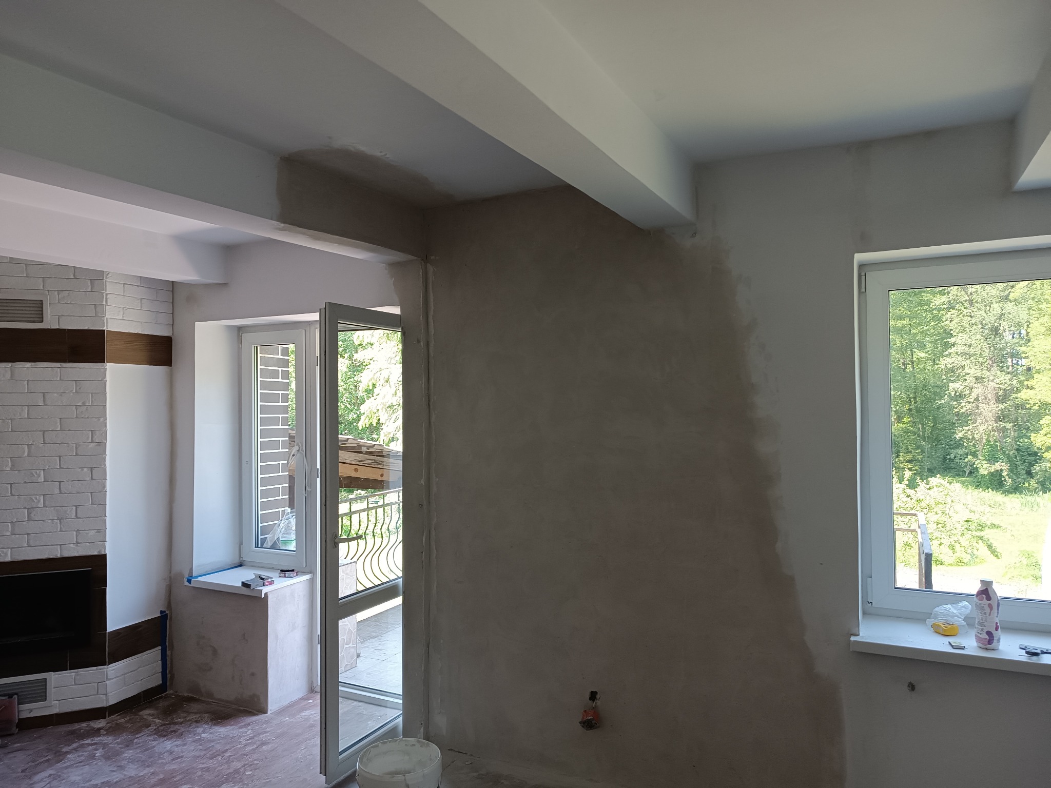 Wnętrze pokoju w trakcie szpachlowania ścian, widoczne częściowo wykończone ściany, okno z widokiem na zieleń, biała stolarka okienna i drzwi balkonowe otwarte na zewnątrz, oraz fragment...