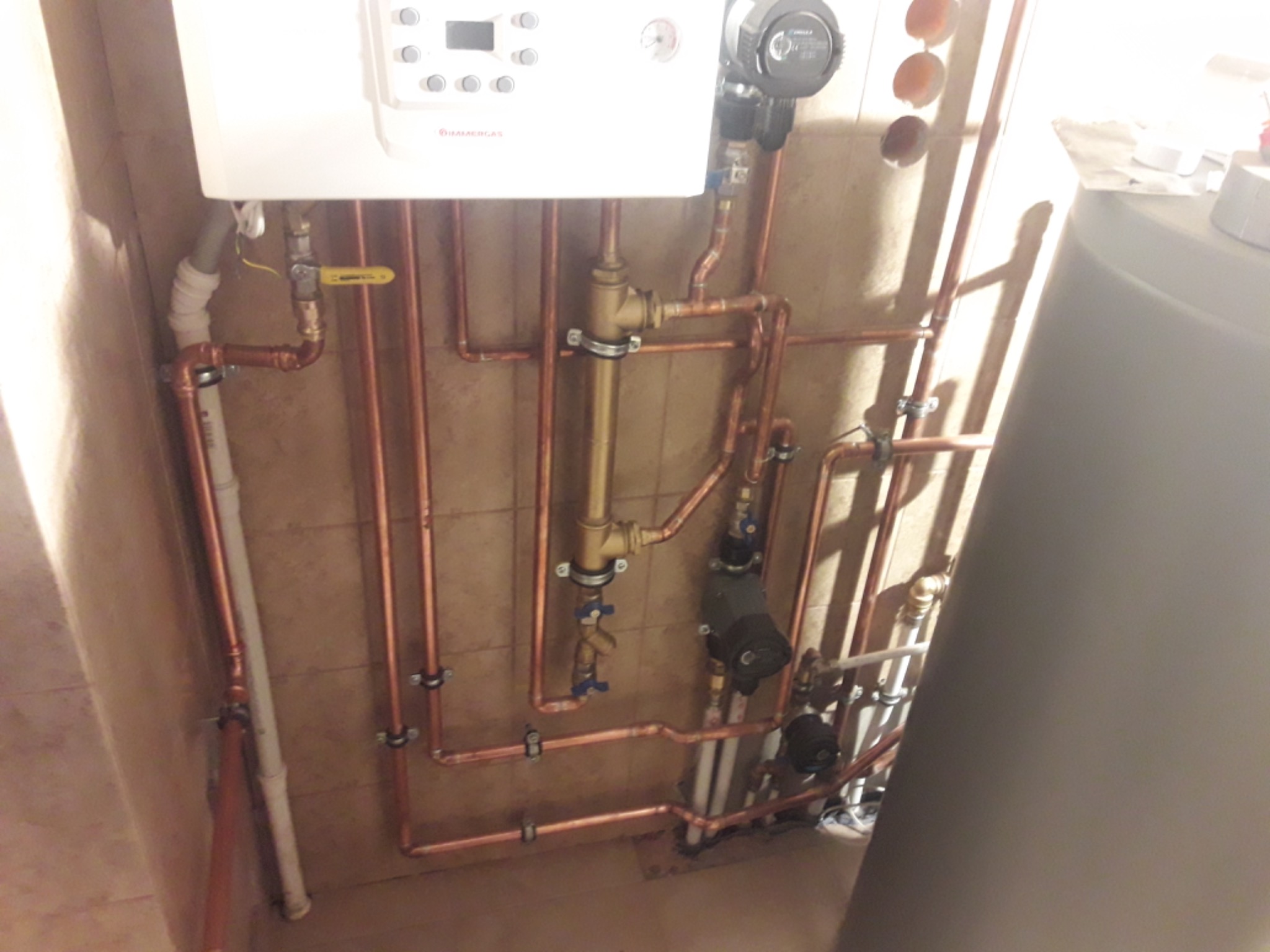 Nowa instalacja grzewcza z miedzianymi rurami i białym kotłem wiszącym na kafelkowanej ścianie, widoczne liczne zawory i elementy montażowe.