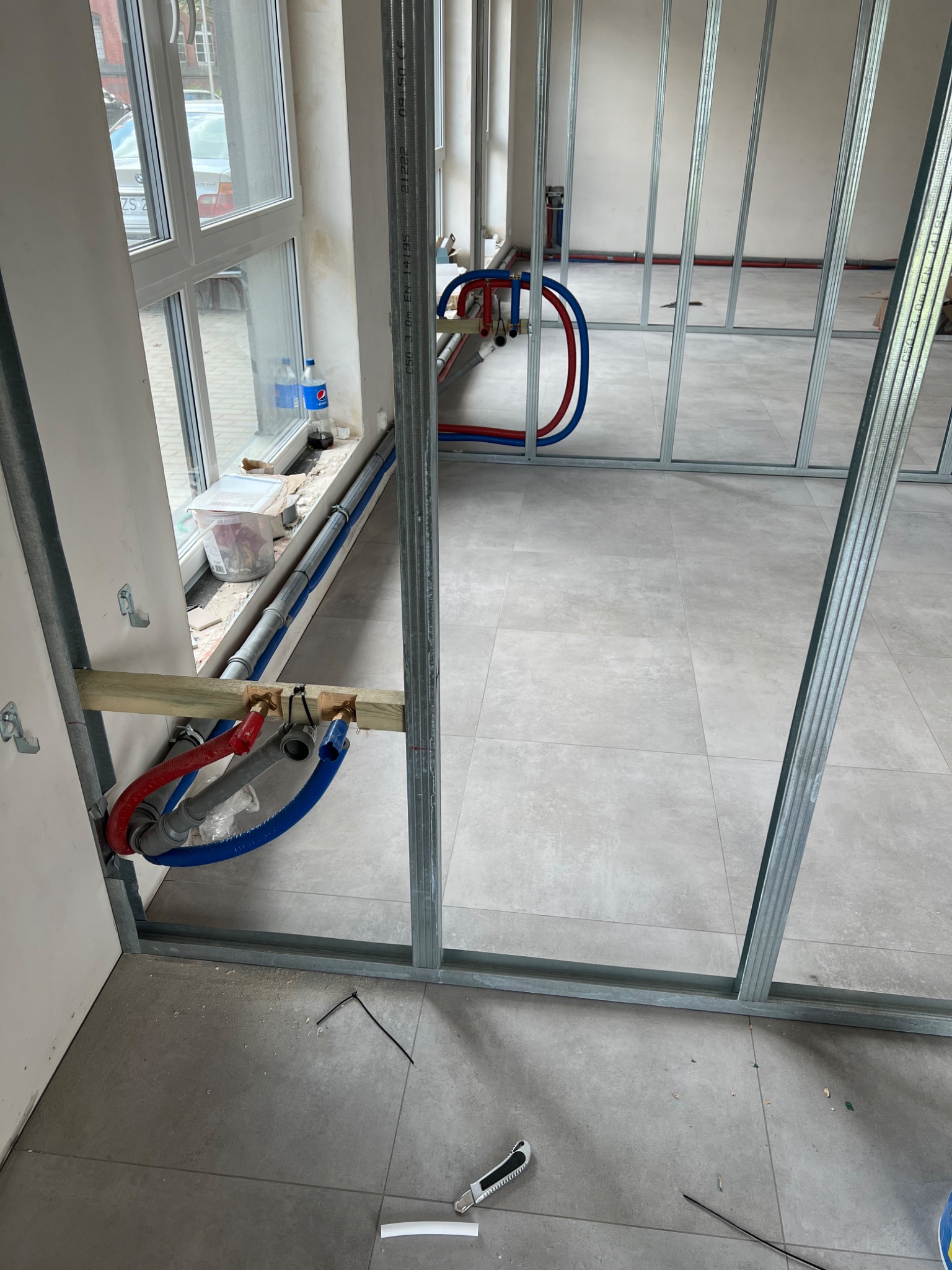 Wnętrze w trakcie remontu z widocznymi, nowo poprowadzonymi rurami instalacji wodnej (czerwone i niebieskie) oraz szarą rurą kanalizacyjną, zamocowanymi do drewnianej belki przy ścianie i pod oknem...