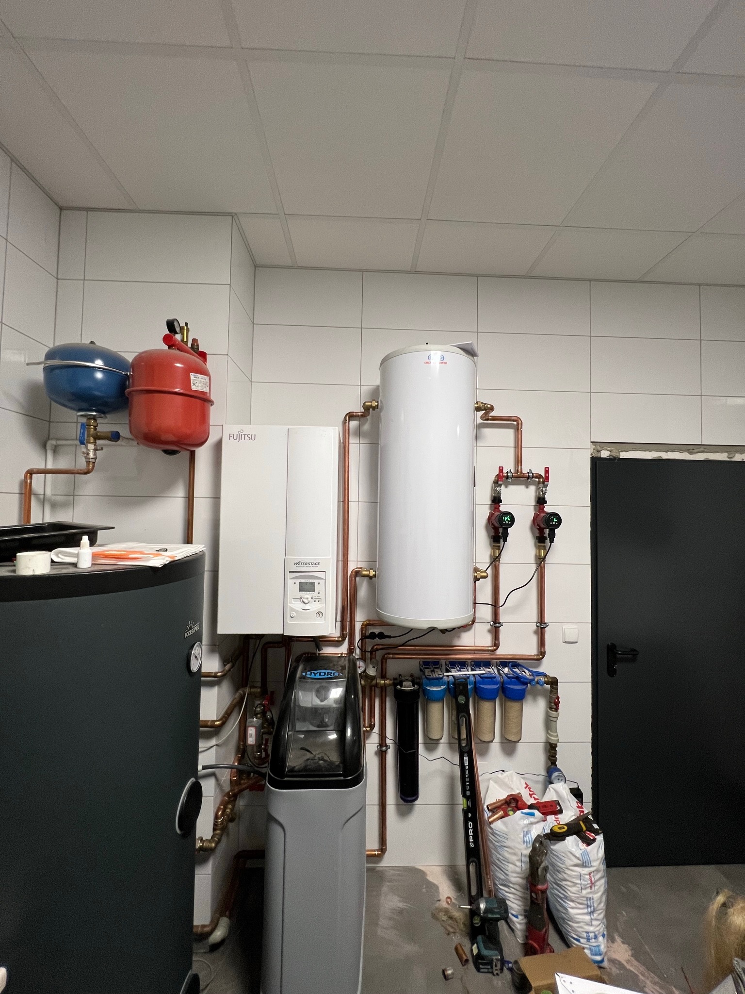 Kompletna kotłownia z piecem gazowym Fujitsu Waterstage, zbiornikiem, zmiękczaczem wody Hydro i filtrami, instalacja z miedzianych rur, worki z materiałami budowlanymi na podłodze.