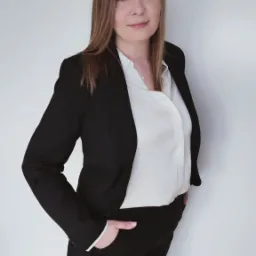 Portret kobiety w czarnym garniturze i białej koszuli, z ręką w kieszeni, prezentującej profesjonalny wizerunek.