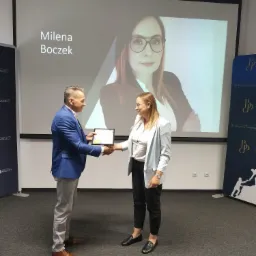 Wręczenie certyfikatu Milenie Boczek na tle jej portretu wyświetlanego na ekranie projektora podczas uroczystości firmowej.