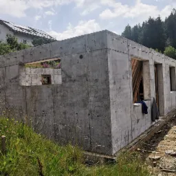 Surowa bryła betonowa w stanie budowy z widocznymi otworami okiennymi, na tle zarośniętego terenu i lasu w oddali. Na ścianie wisi kurtka i koszula w kratę.