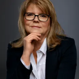 Portret kobiety w okularach i ciemnej marynarce, opierającej podbródek na dłoni, na szarym tle.