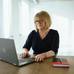 Kobieta w okularach, w biurowym wnętrzu, pracuje na laptopie, obok leży notatnik z długopisem. Widoczne białe plamy na skórze dłoni.