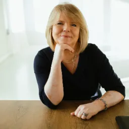 Portret uśmiechniętej kobiety z blond włosami, opierającej brodę na dłoni, siedzącej za drewnianym biurkiem w jasnym biurze.