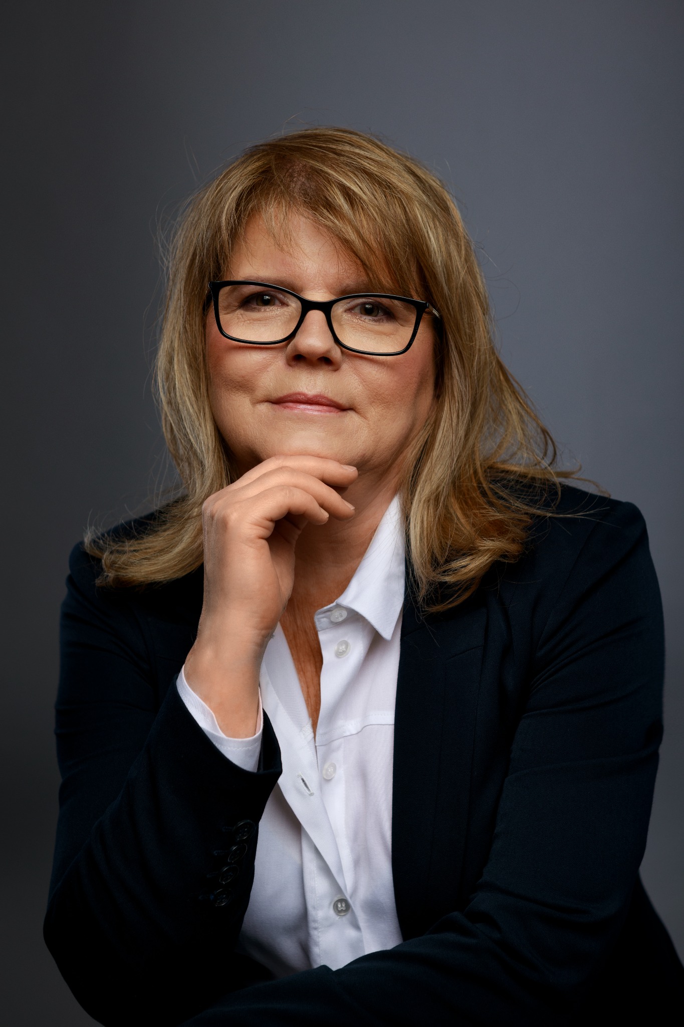 Portret kobiety w okularach i ciemnej marynarce, opierającej podbródek na dłoni, na szarym tle.