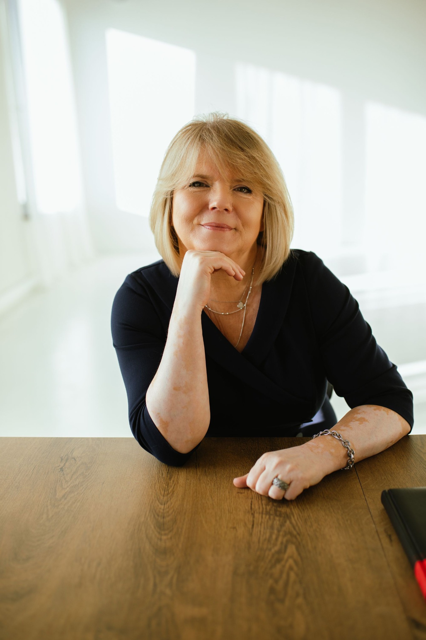 Portret uśmiechniętej kobiety z blond włosami, opierającej brodę na dłoni, siedzącej za drewnianym biurkiem w jasnym biurze.