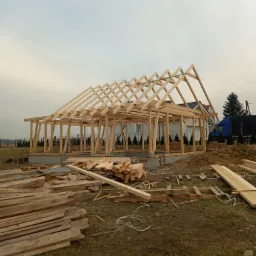 Konstrukcja drewniana domu w trakcie budowy, widoczna więźba dachowa i fundamenty, na pierwszym planie stos drewna budowlanego, pochmurne niebo w tle.