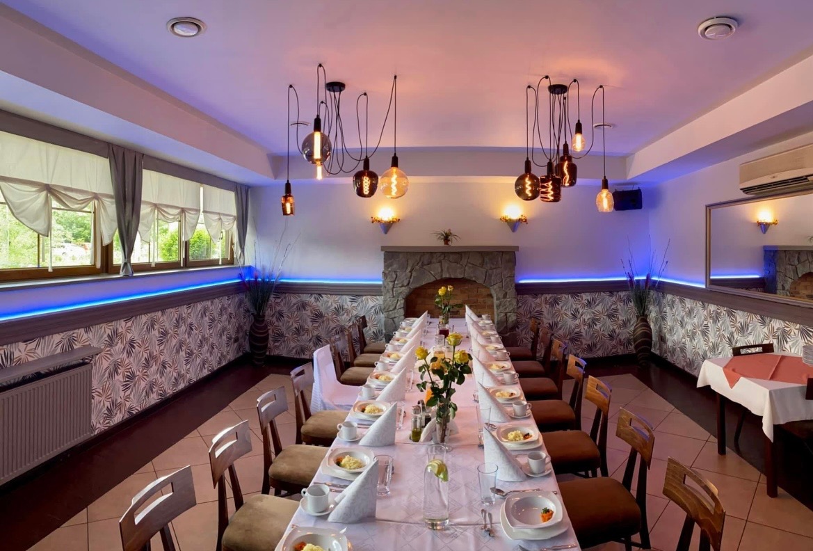 Długi stół nakryty białym obrusem z zastawą i dekoracjami kwiatowymi, przygotowany dla gości w sali z kamiennym kominkiem i nowoczesnym oświetleniem.