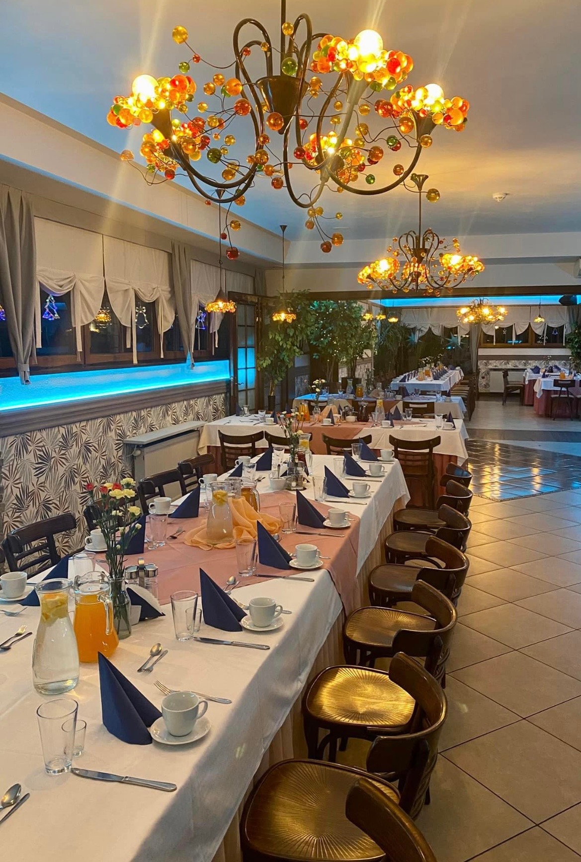 Uroczysta sala bankietowa z długim stołem nakrytym białym obrusem i różowym bieżnikiem, dekoracje z ciemnoniebieskich serwetek, szklanek z lemoniadą i kwiatów, nad stołem ozdobne żyrandole...