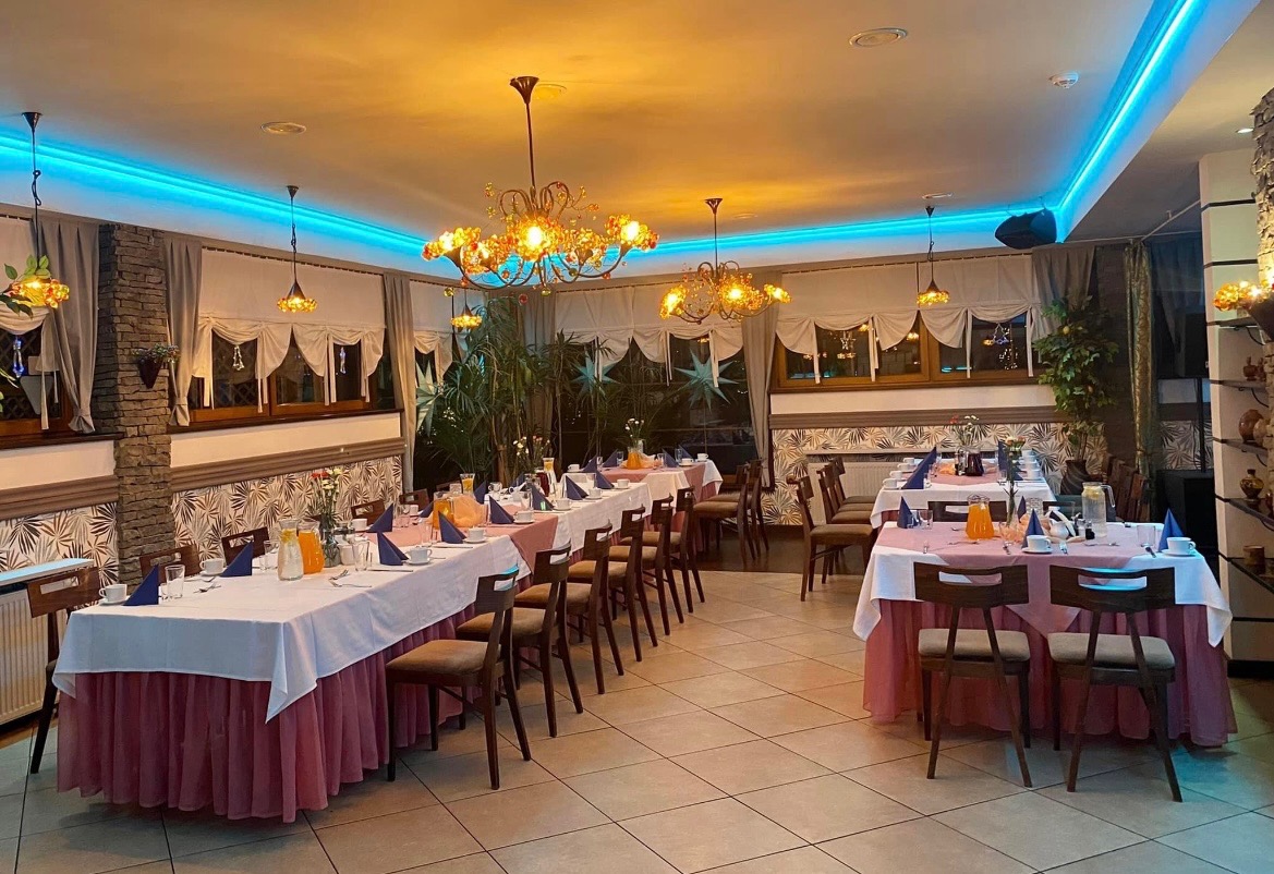 Uroczysta sala restauracyjna z nakrytymi stołami, białe obrusy z różowym akcentem, niebieskie oświetlenie LED na suficie, żyrandole w stylu retro, przygotowana na przyjęcie.