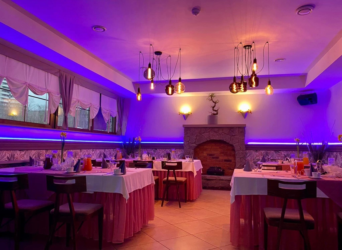 Uroczysta sala restauracyjna z nakrytymi stołami, oświetlona fioletowym światłem, z dekoracyjnym kominkiem i wiszącymi lampami w stylu retro.