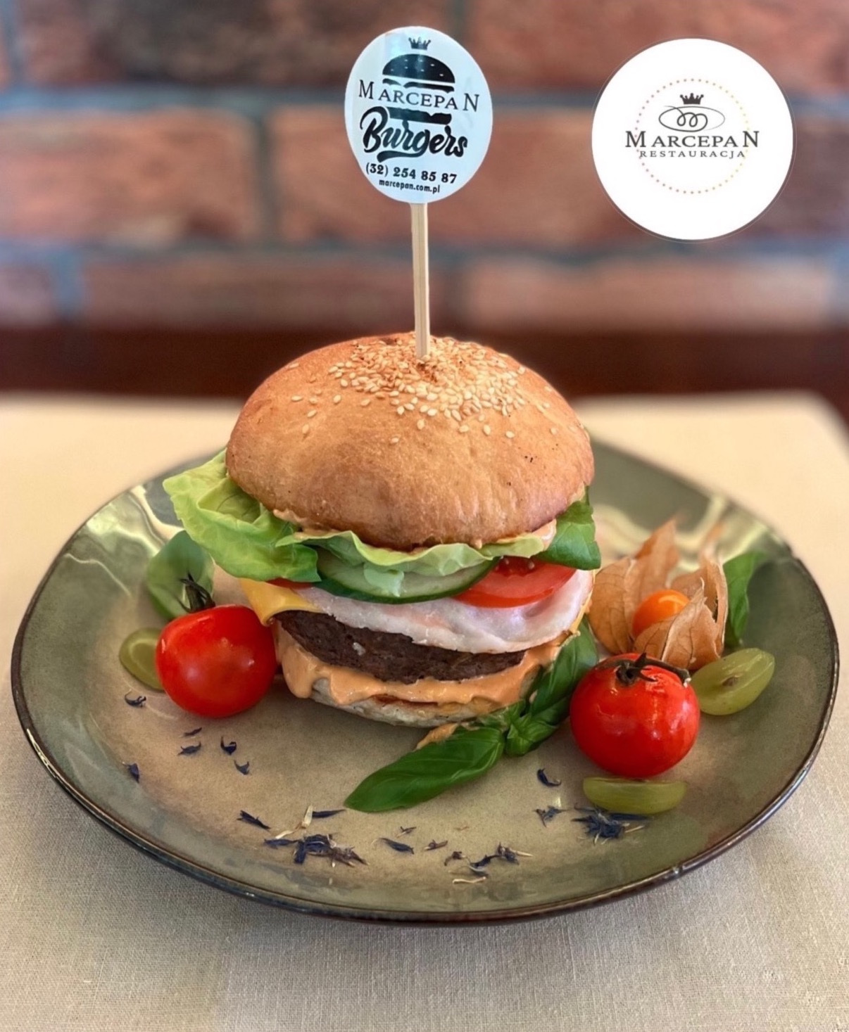 Apetyczny burger na ceramicznym talerzu, udekorowany pomidorkami cherry, physalis i bazylią, z logo Marcepan Burgers wbitym w górną bułkę.