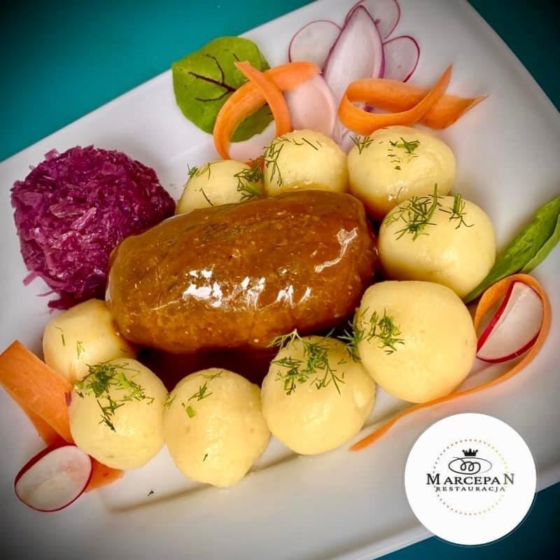 Elegancko podane danie główne: pieczeń w towarzystwie ziemniaków posypanych koperkiem, czerwonej kapusty i dekoracji z rzodkiewki i marchewki, talerz z logiem restauracji Marcepan.