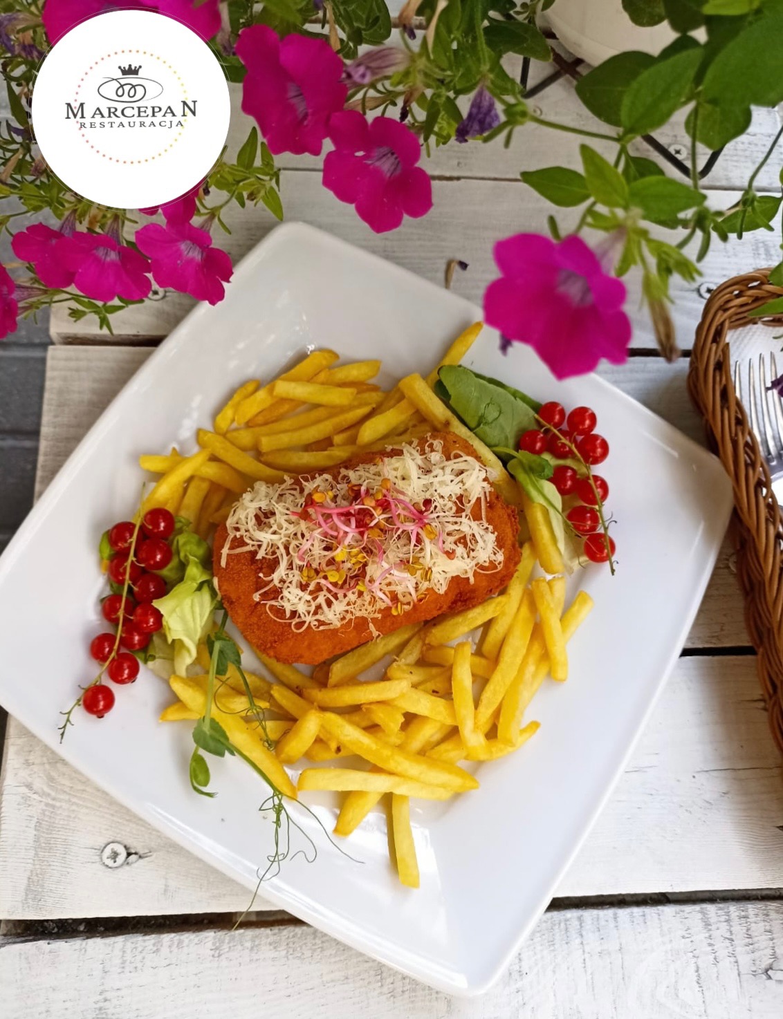 Kotlet schabowy z frytkami, posypany serem i kiełkami, udekorowany czerwoną porzeczką i sałatą, podany na białym kwadratowym talerzu z kwiatami w tle.