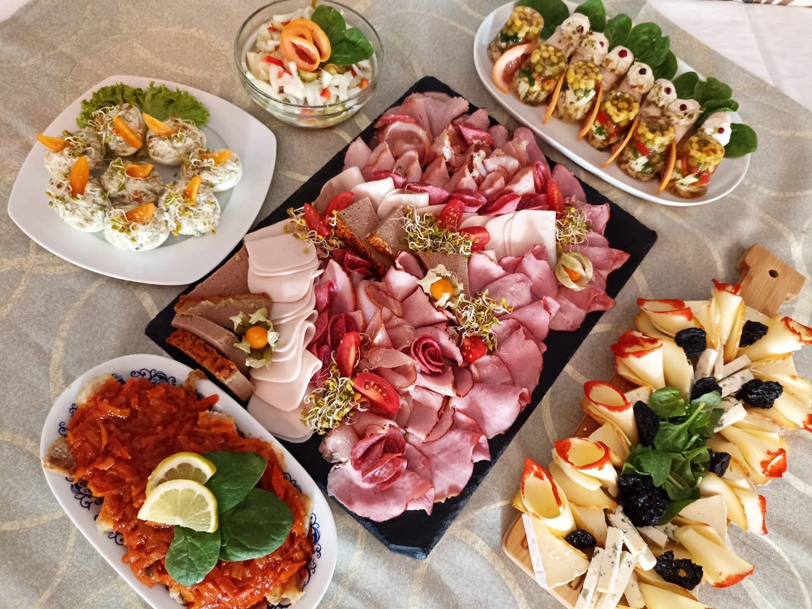 Dekoracyjny stół cateringowy z różnorodnymi przekąskami: wędliny na czarnej desce, jajka faszerowane, sałatka warzywna w szklanej misce, roladki z farszem na talerzu, leczo z cytryną i deska serów...