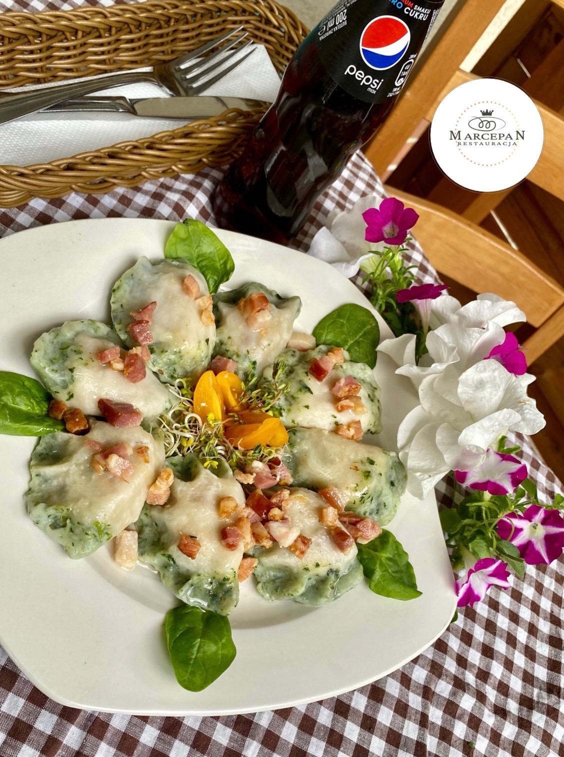 Porcja zielonych pierogów okraszonych boczkiem, podanych na białym talerzu z dekoracją z kwiatów i ziół, obok butelka Pepsi i koszyk z sztućcami na brązowo-białej kratowanej serwecie.
