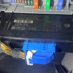 Skrzynka bezpieczników samochodowych z widocznymi kolorowymi bezpiecznikami i niebieskim złączem elektrycznym, widoczne kable i śruby.