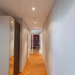 Długi korytarz z parkietem ułożonym w jodełkę, oświetlony punktowymi lampami w suficie, z lustrzanymi drzwiami szafy po lewej stronie i widokiem na dalsze pomieszczenia przez przeszklone drzwi...