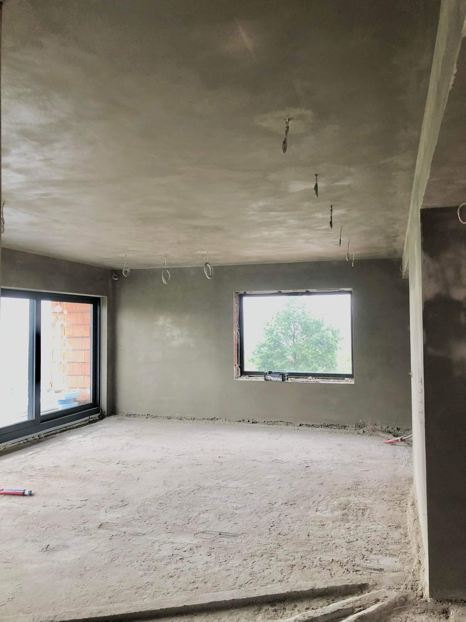 Wnętrze pokoju po nałożeniu gładzi cementowej na ściany i sufit, widoczne wystające przewody elektryczne, okno z widokiem na drzewo i częściowo zakryte okno balkonowe.