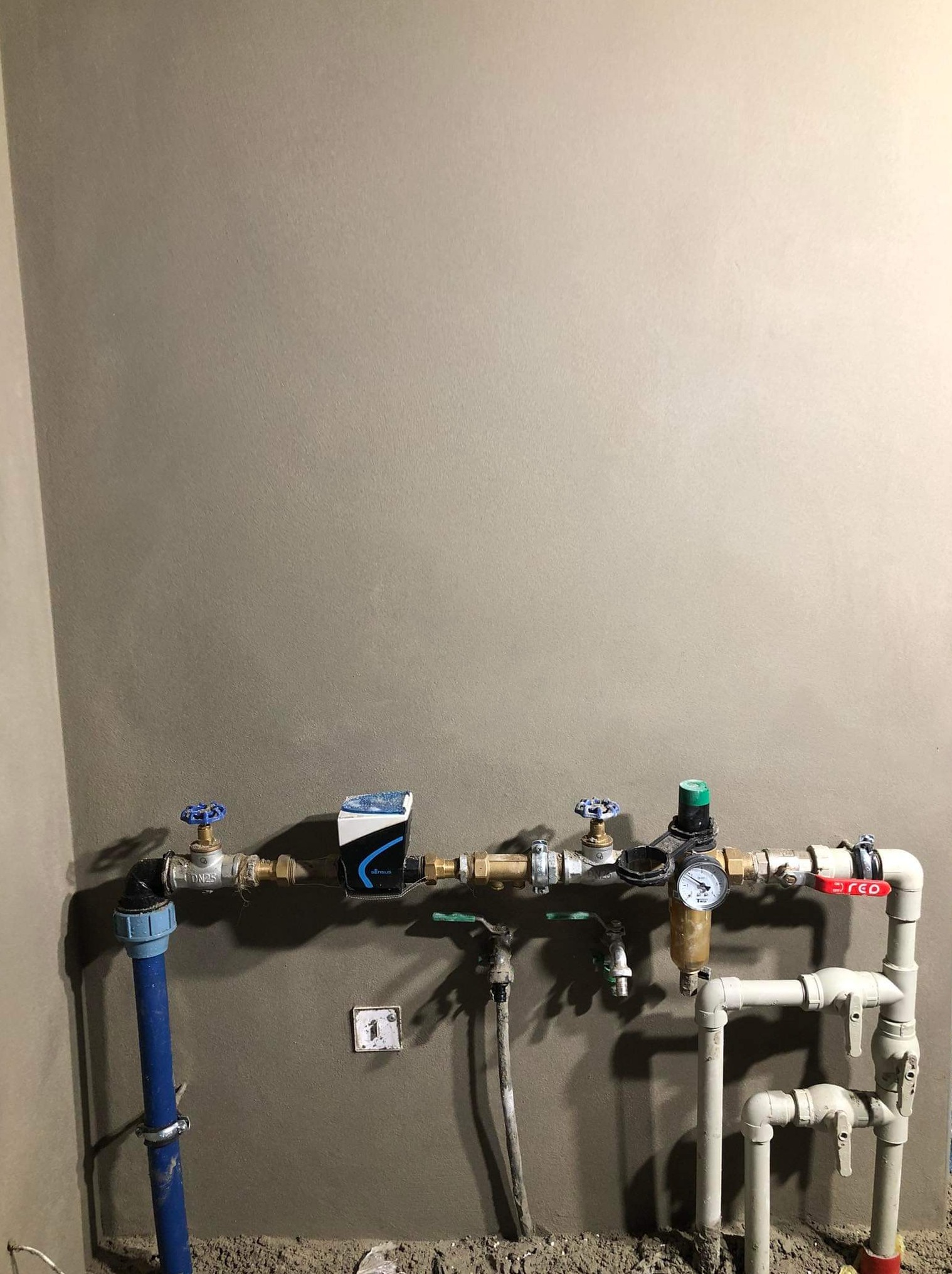 Świeżo otynkowana ściana z zamontowaną instalacją wodną: rury PCV, zawory z niebieskimi pokrętłami, wodomierz Sensus i manometr.
