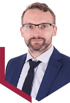 Portret mężczyzny w okularach, ciemnym garniturze i krawacie na białym tle z bordowym elementem graficznym po lewej stronie.