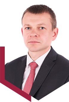 Portret mężczyzny w garniturze i czerwonym krawacie na białym tle, z bordowym elementem graficznym w lewym dolnym rogu.