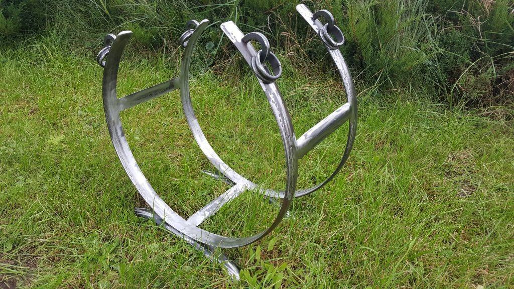 Dwa metalowe, spawane półkola z poprzeczkami i ozdobnymi pierścieniami, prawdopodobnie elementy balustrady, leżą na trawie.
