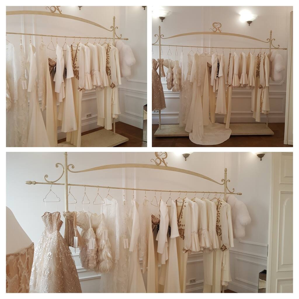 Kremowe suknie i żakiety ślubne wiszące na ozdobnym, metalowym wieszaku w butiku z białymi ścianami i sztukaterią.