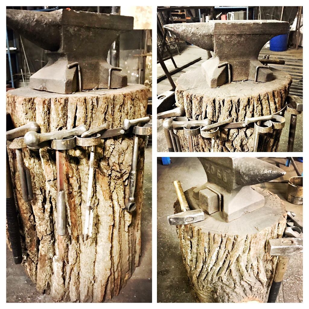 Kowadło na pniu drzewa z zawieszonymi narzędziami kowalskimi, widoczne różne młotki i stemple, w tle warsztat.