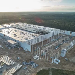 Szeroki kadr z lotu ptaka na budowę hali magazynowej z częściowo ukończonym dachem, widoczne materiały budowlane, konstrukcja stalowa i otaczający las w promieniach zachodzącego słońca.