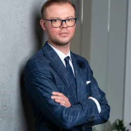 Portret prawnika w niebieskim garniturze w kratę, stojącego na tle betonowej ściany i roślin doniczkowych.