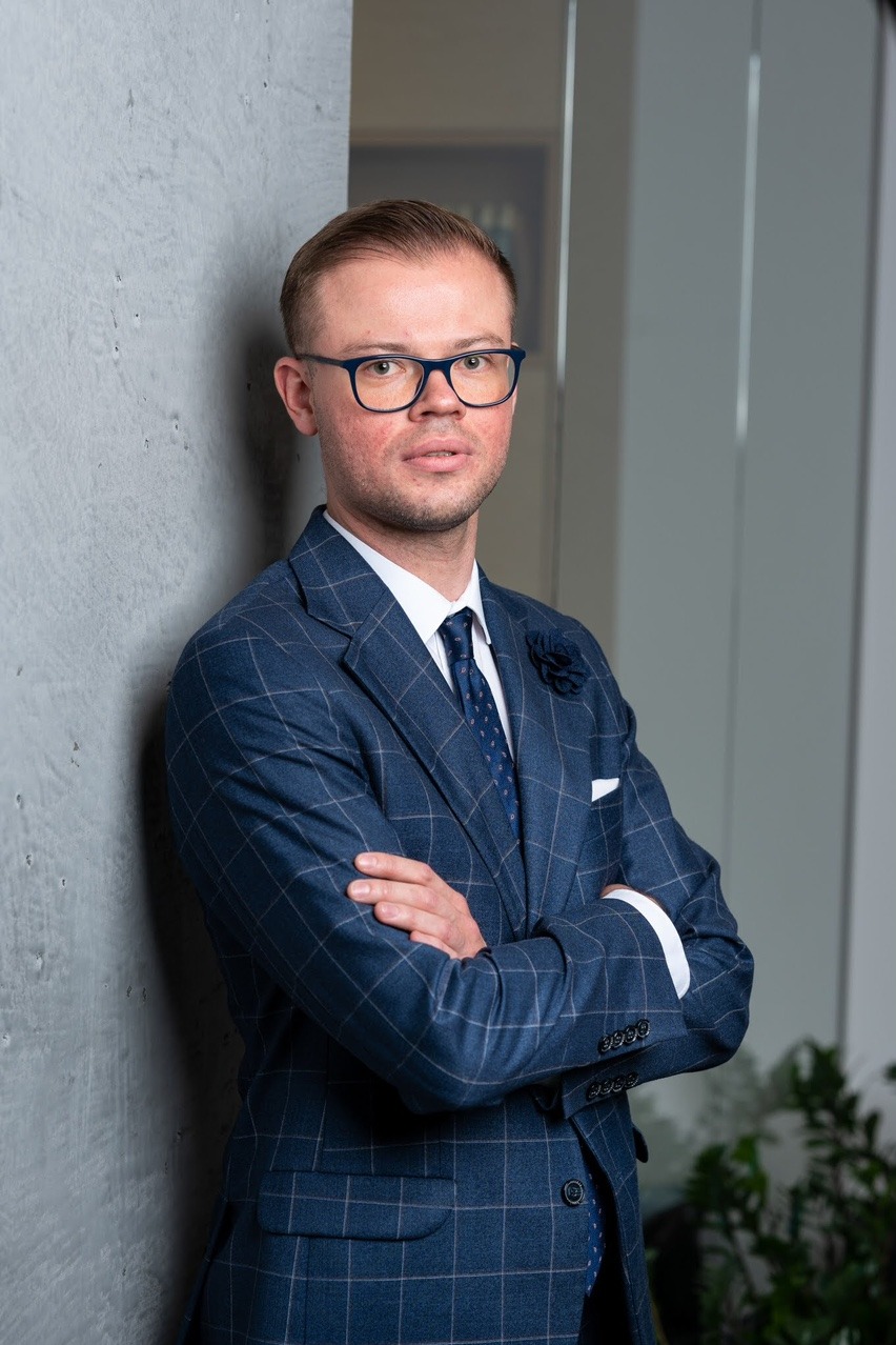 Portret prawnika w niebieskim garniturze w kratę, stojącego na tle betonowej ściany i roślin doniczkowych.