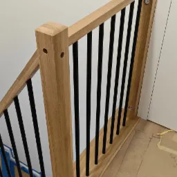 Fragment drewnianej balustrady schodowej z czarnymi, pionowymi elementami. Jasne drewno, minimalistyczny design, widoczne mocowania.