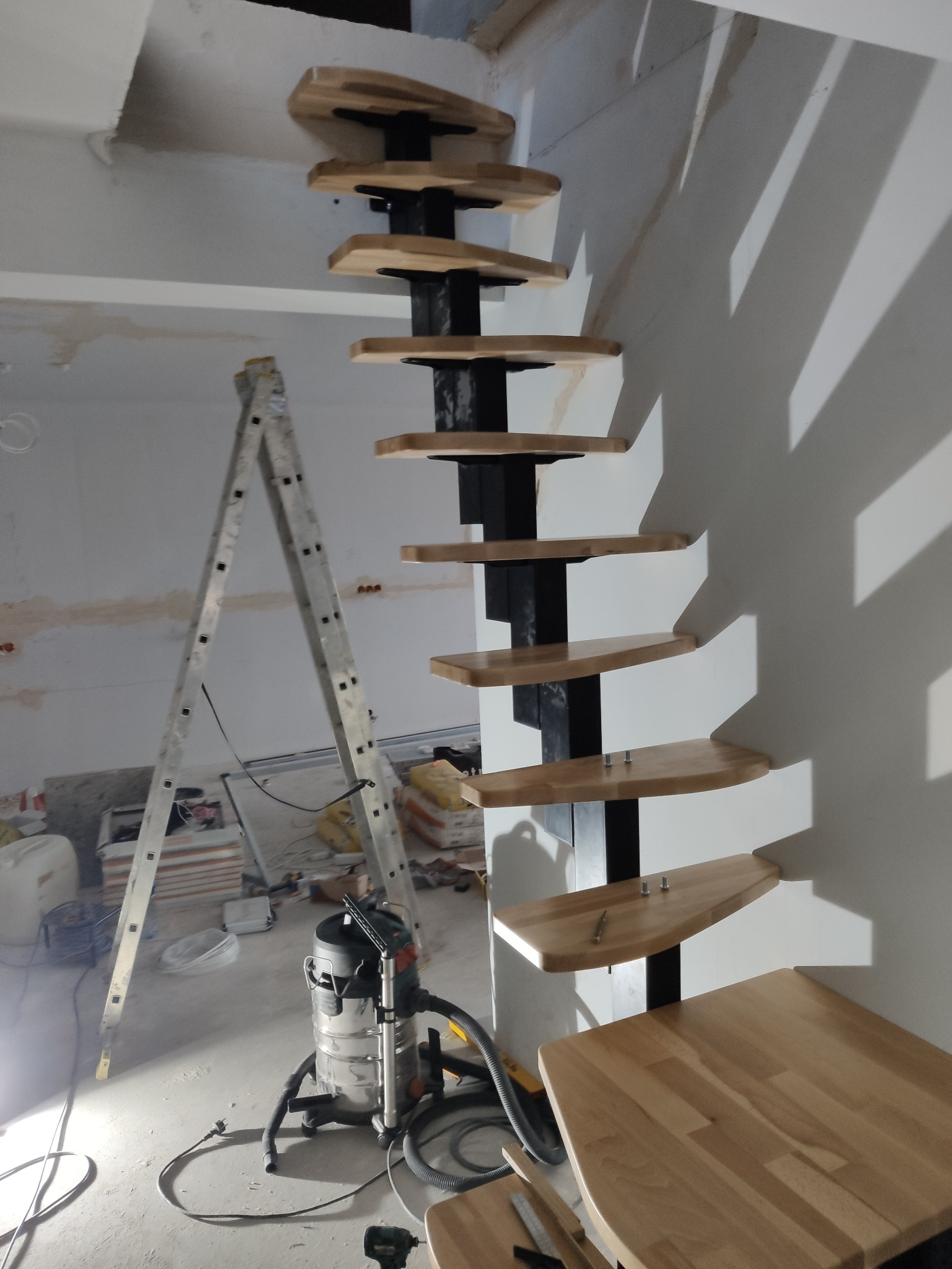 Nowoczesne schody spiralne z drewnianymi stopniami i czarną metalową konstrukcją, w trakcie montażu w surowym wnętrzu z drabiną i narzędziami.