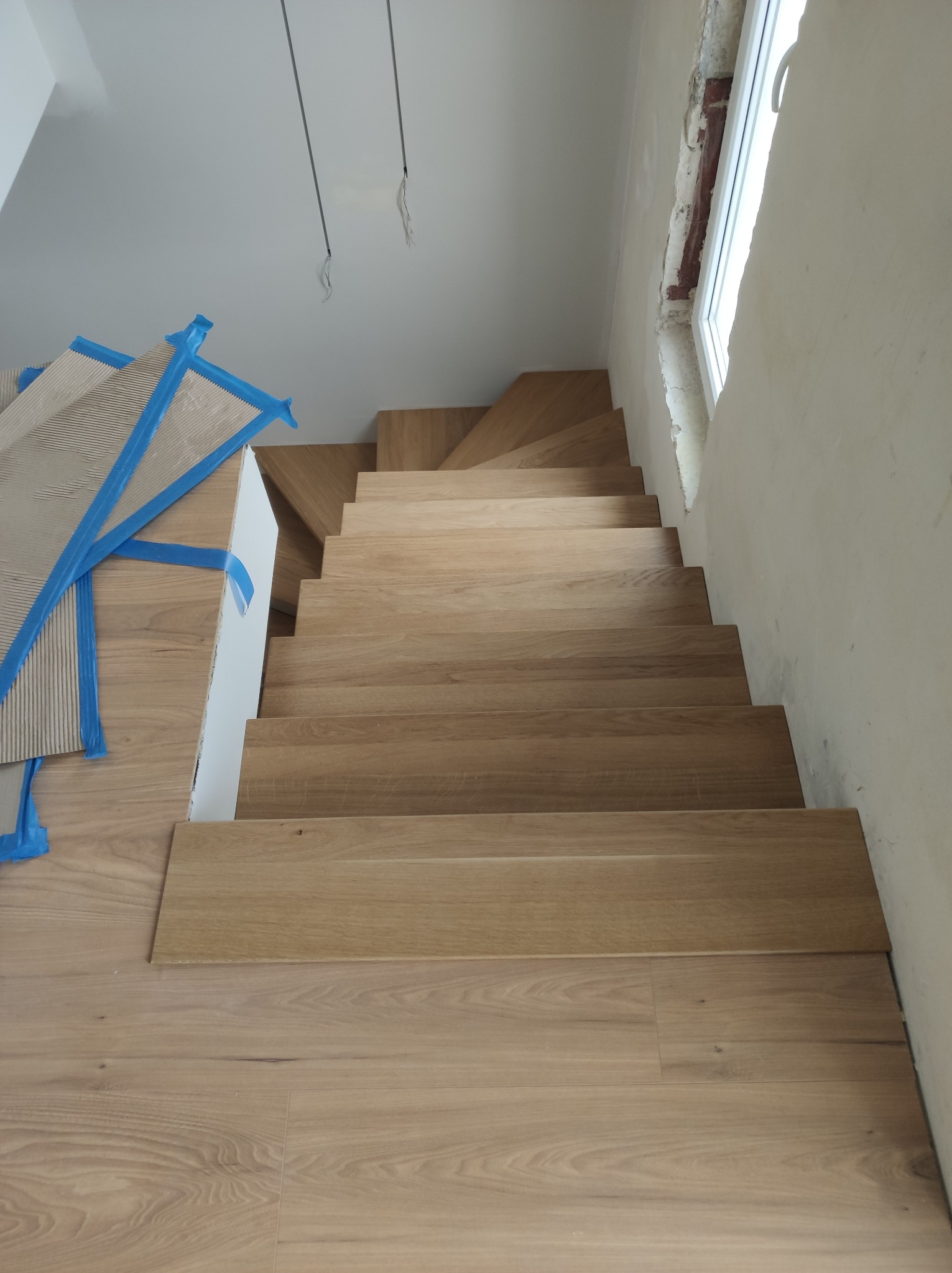 Montaż drewnianych schodów z widocznymi elementami wykończeniowymi zabezpieczonymi taśmą. Widok z góry na surowe ściany i fragment okna.