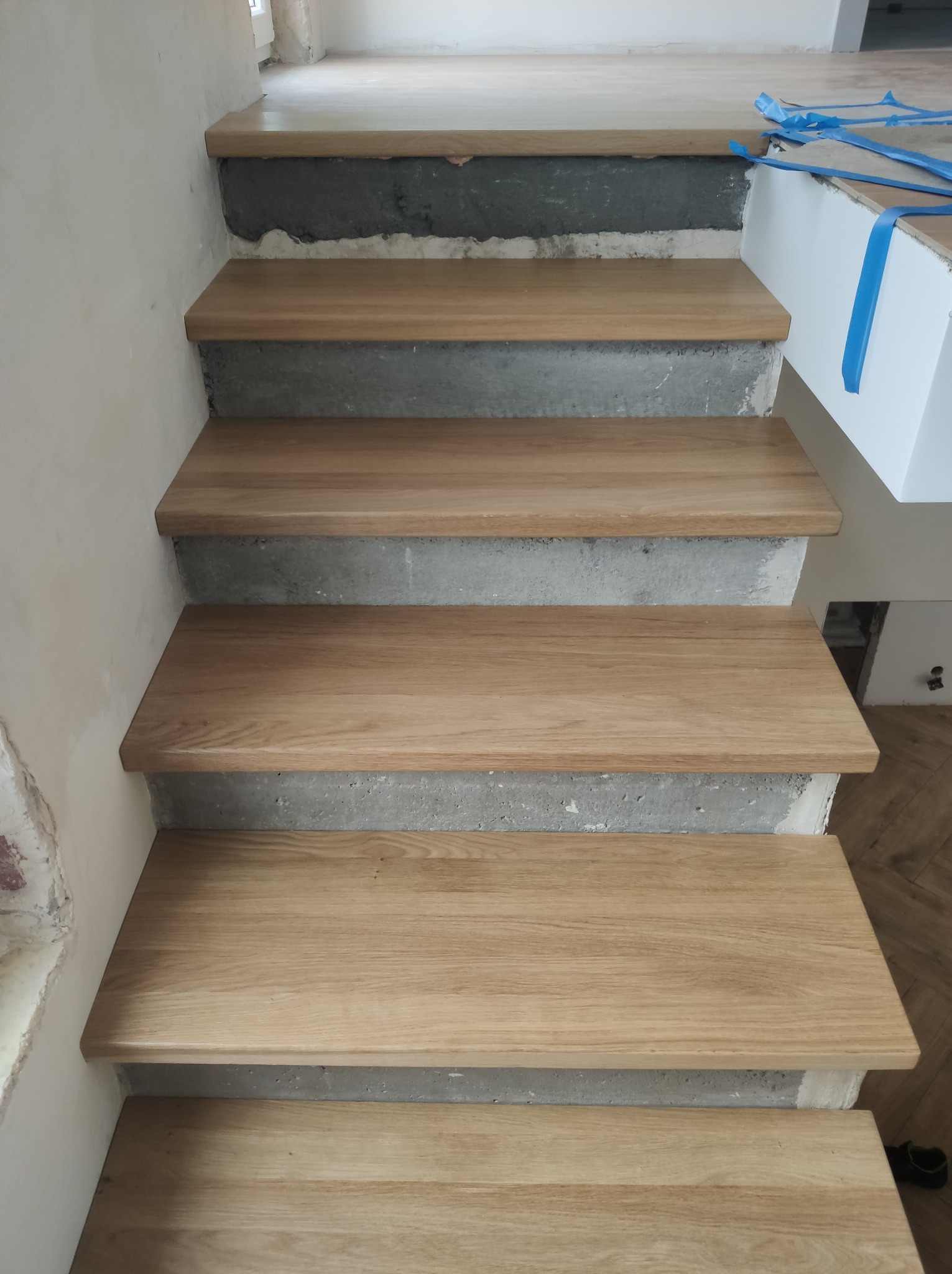 Drewniane stopnie schodów z betonu, widok z góry. Jasne drewno, surowy beton. Fragment wnętrza w trakcie wykańczania.