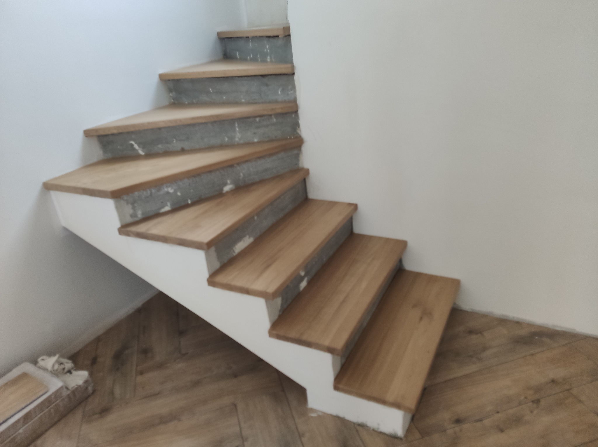 Drewniane schody w trakcie montażu, z widocznymi betonowymi stopniami i drewnianymi nakładkami. Podłoga wyłożona parkietem w jodełkę.