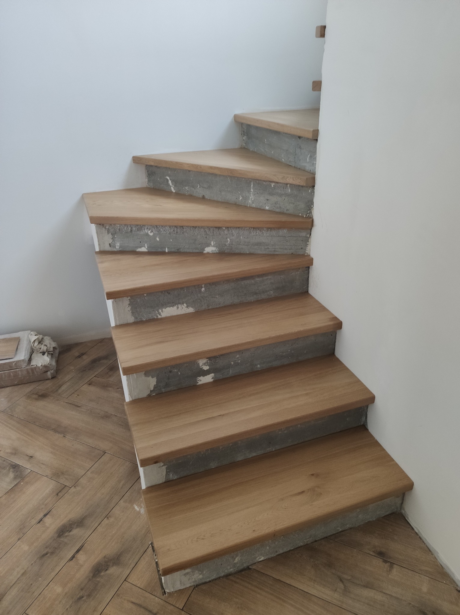Drewniane stopnie schodów z surowym betonem, ułożone na jasnej podłodze w jodełkę, przy białej ścianie. Widoczne niedokończone wykończenie.