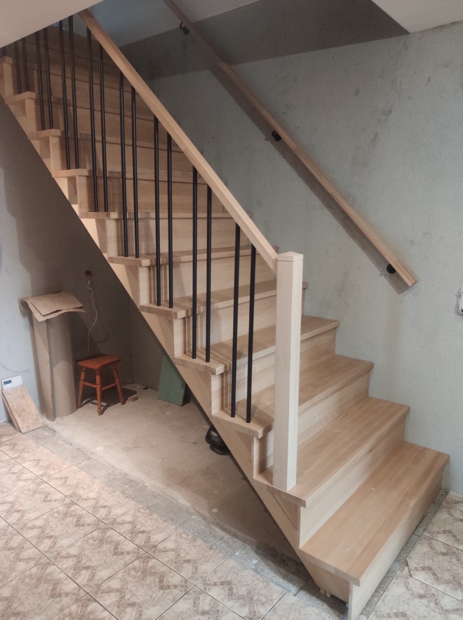 Jasne, drewniane schody z czarnymi balustradami w surowym wnętrzu. Widoczna poręcz zamontowana na ścianie. Podłoga wyłożona płytkami.