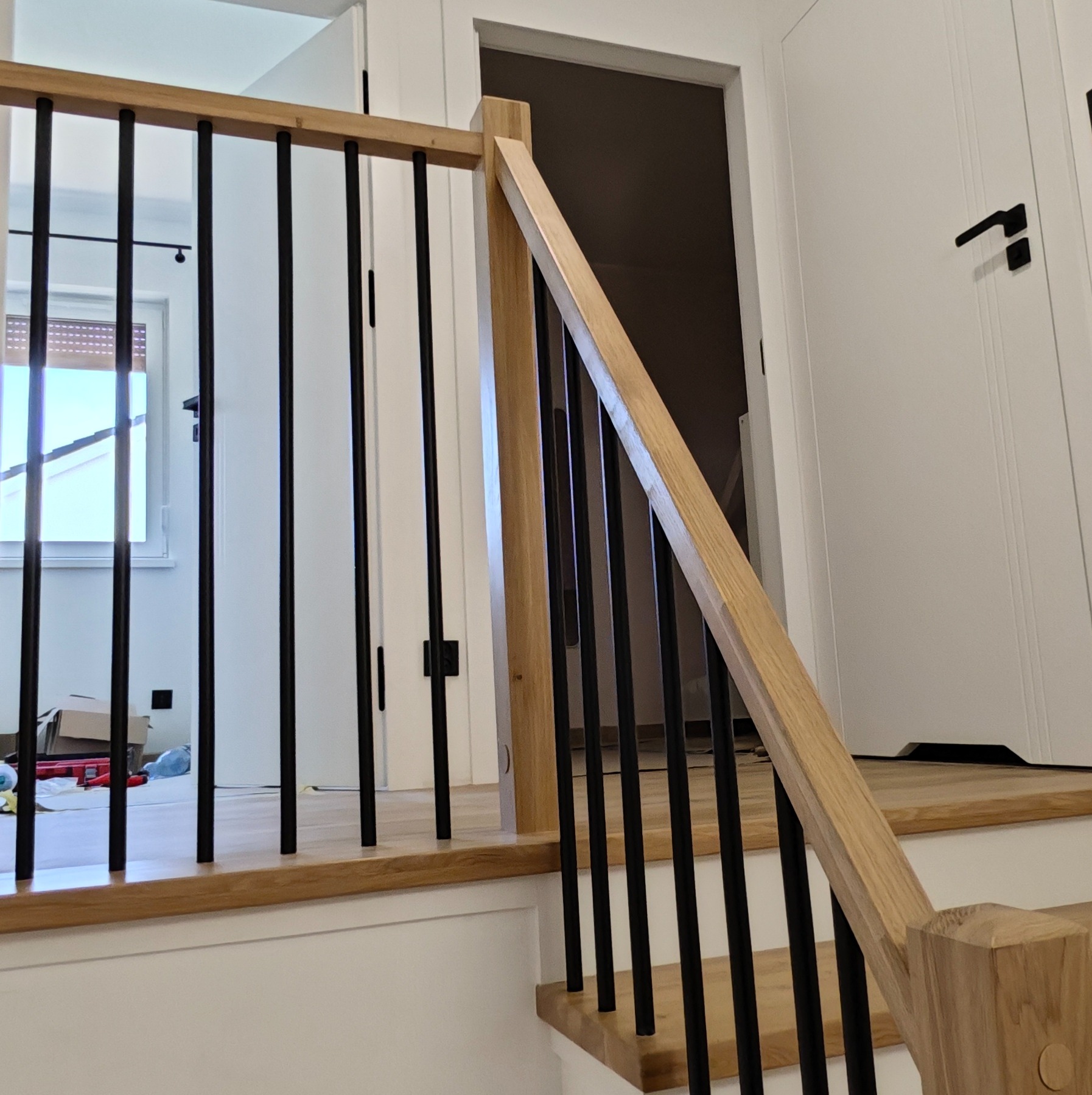 Drewniane schody z czarną, metalową balustradą w nowoczesnym wnętrzu. Widoczne białe drzwi i fragment okna z roletą. Jasne drewno kontrastuje z czarnymi elementami.