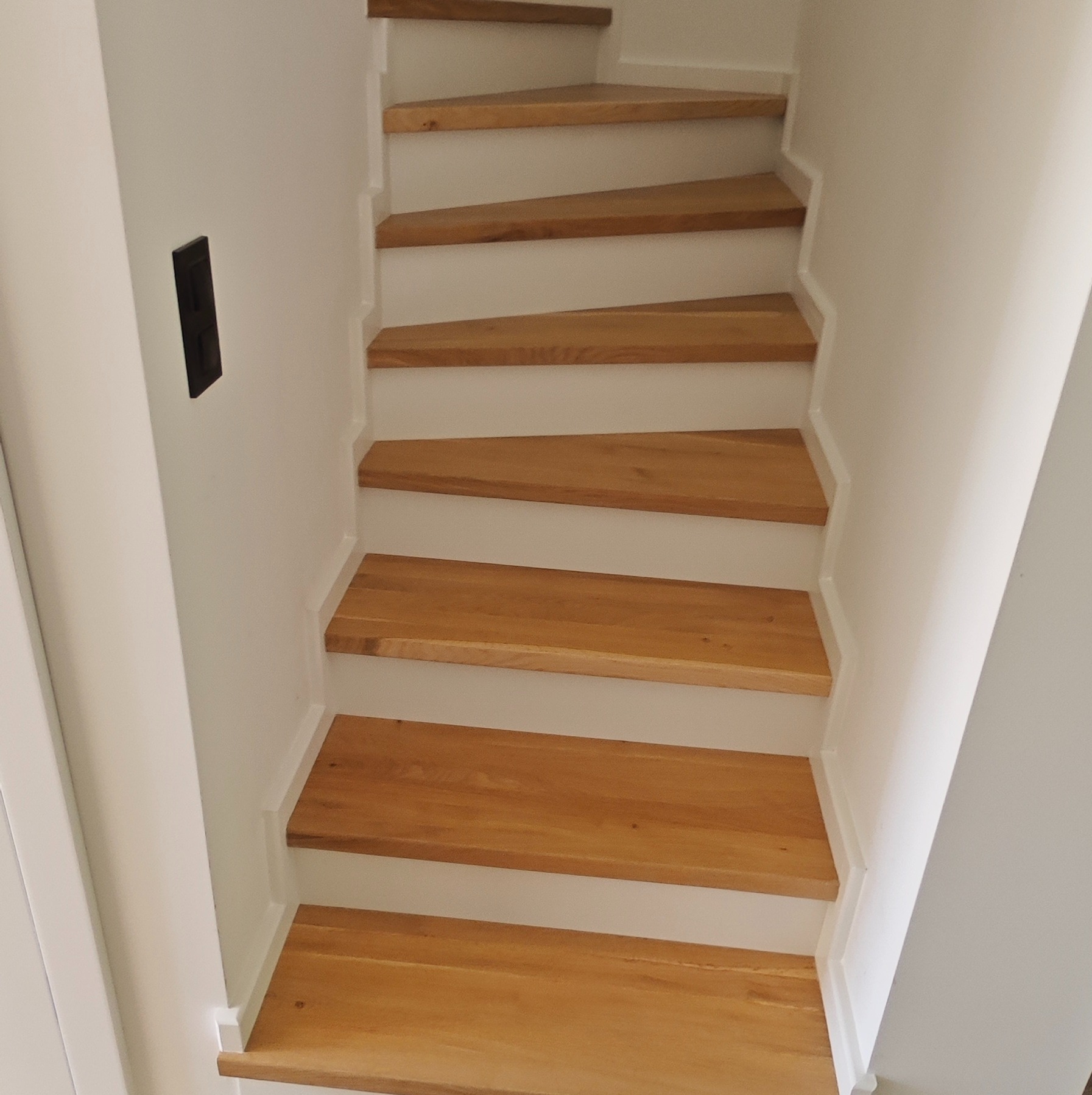 Drewniane schody o jasnym odcieniu drewna, z białymi elementami wykończeniowymi, widziane z dołu, z czarnym włącznikiem światła na ścianie.
