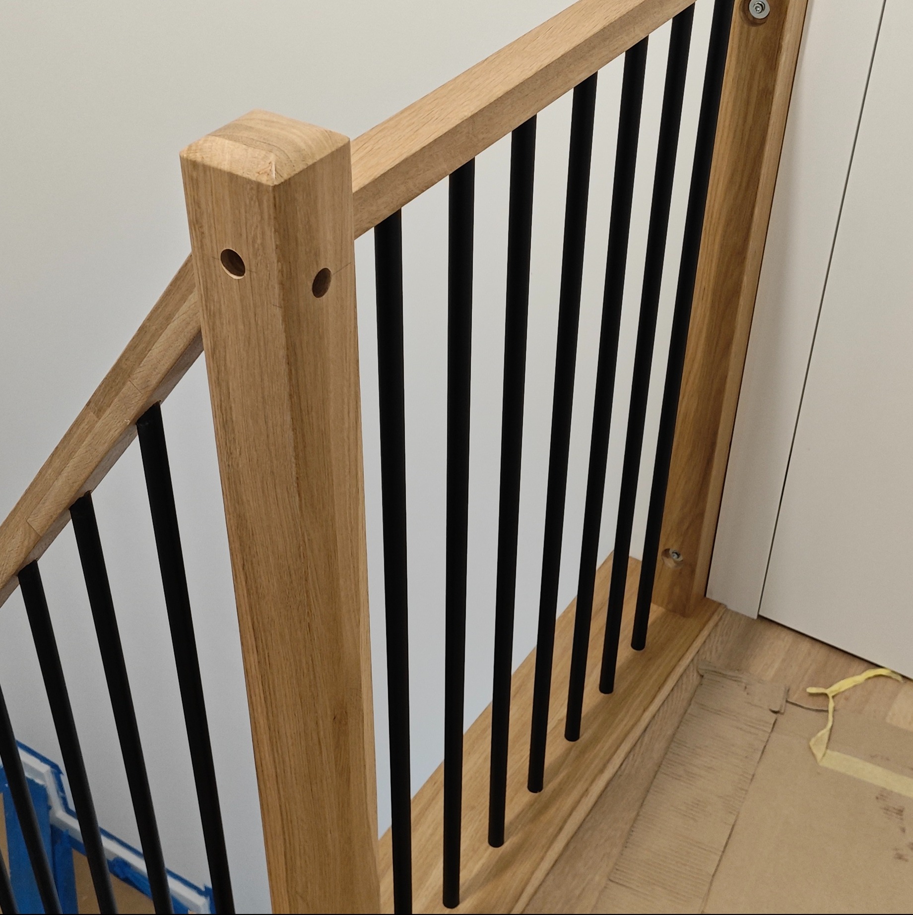 Fragment drewnianej balustrady schodowej z czarnymi, pionowymi elementami. Jasne drewno, minimalistyczny design, widoczne mocowania.