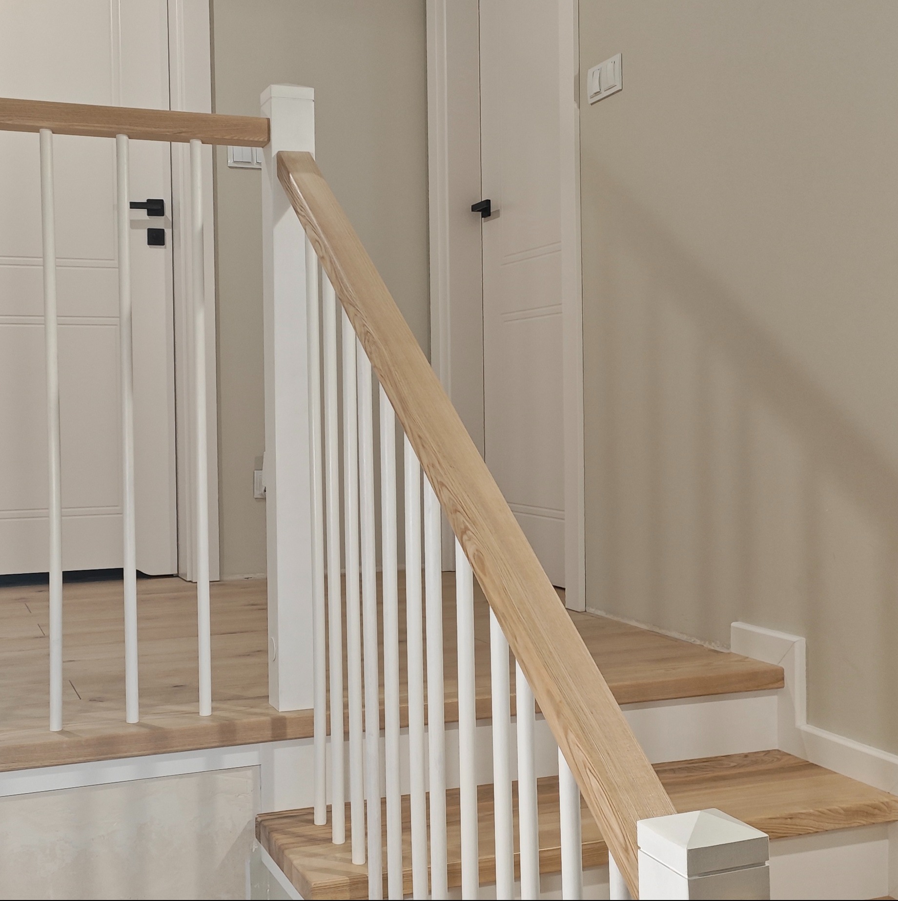 Drewniane schody z białą balustradą w nowoczesnym wnętrzu. Jasne drewno stopni kontrastuje z bielą poręczy i tralek. W tle widoczne białe drzwi.